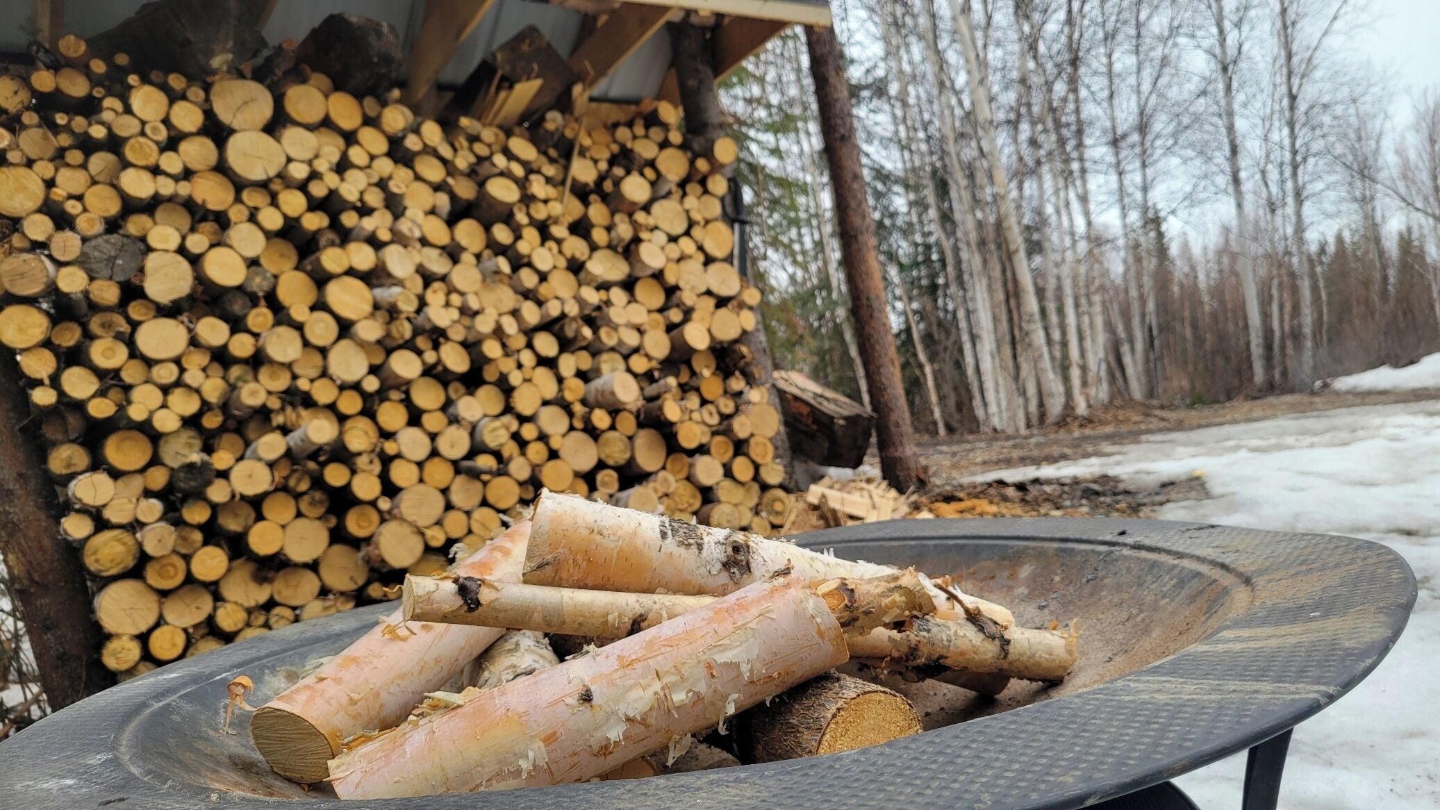 Ein metallener Feuerkorb, gefüllt mit Holzscheiten, ein Stapel geschnittenen Holzes im Hintergrund und eine verschneite Landschaft mit Bäumen.