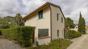 Una casa pequeña de un piso, con fachada color beige y contraventanas verdes, rodeada por un camino de grava y un jardín con flores rosas.
