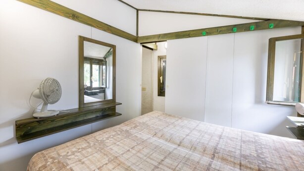 Un dormitorio con cama, un ventilador de noche, un espejo y techo con vigas de madera.