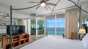 A hotel room with a large bed, a TV on a wooden stand, a ceiling fan, and a balcony with a view of the ocean.