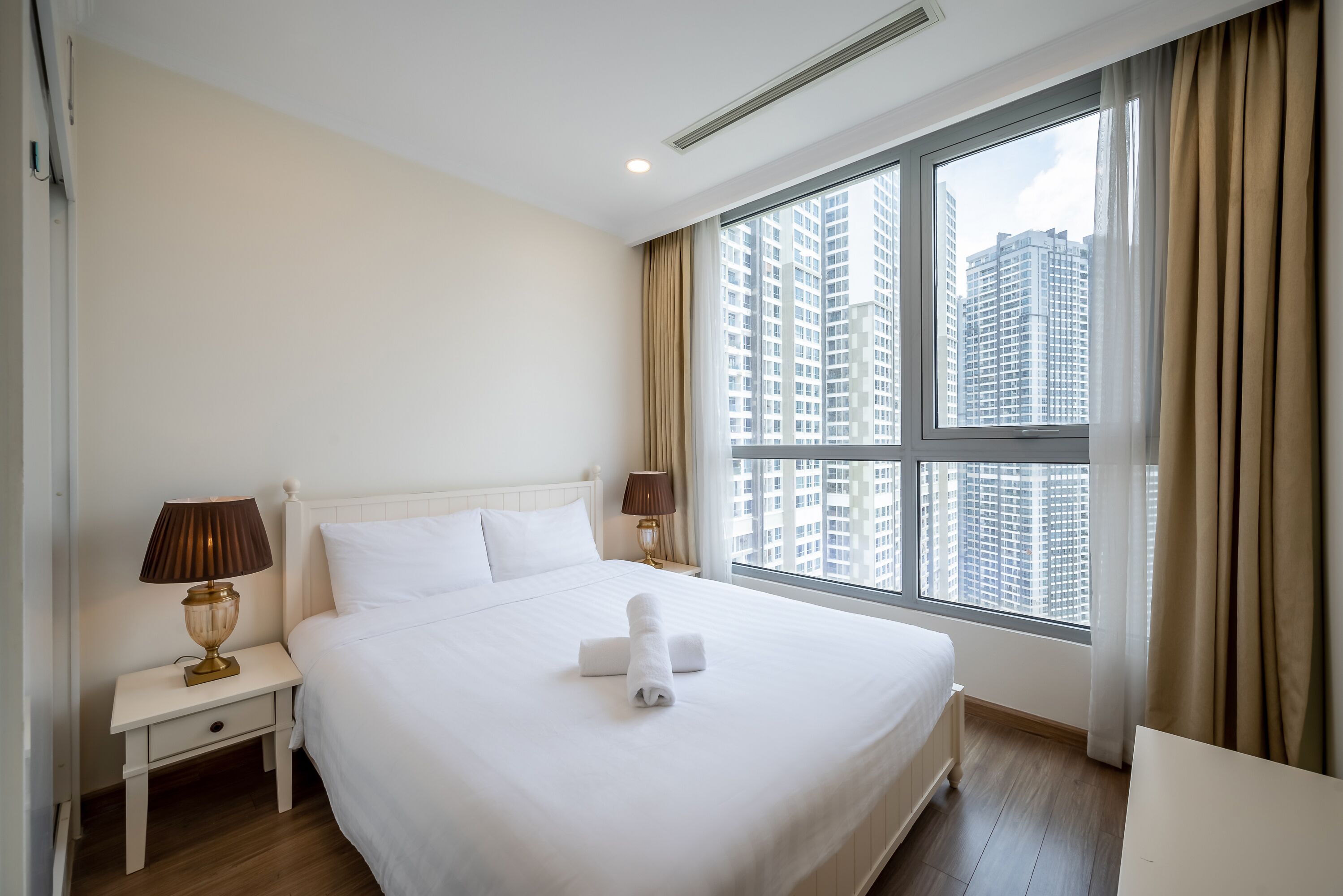 Una habitación de hotel con una cama grande, dos mesitas de noche con lámparas, una ventana con cortinas y vistas a edificios altos.