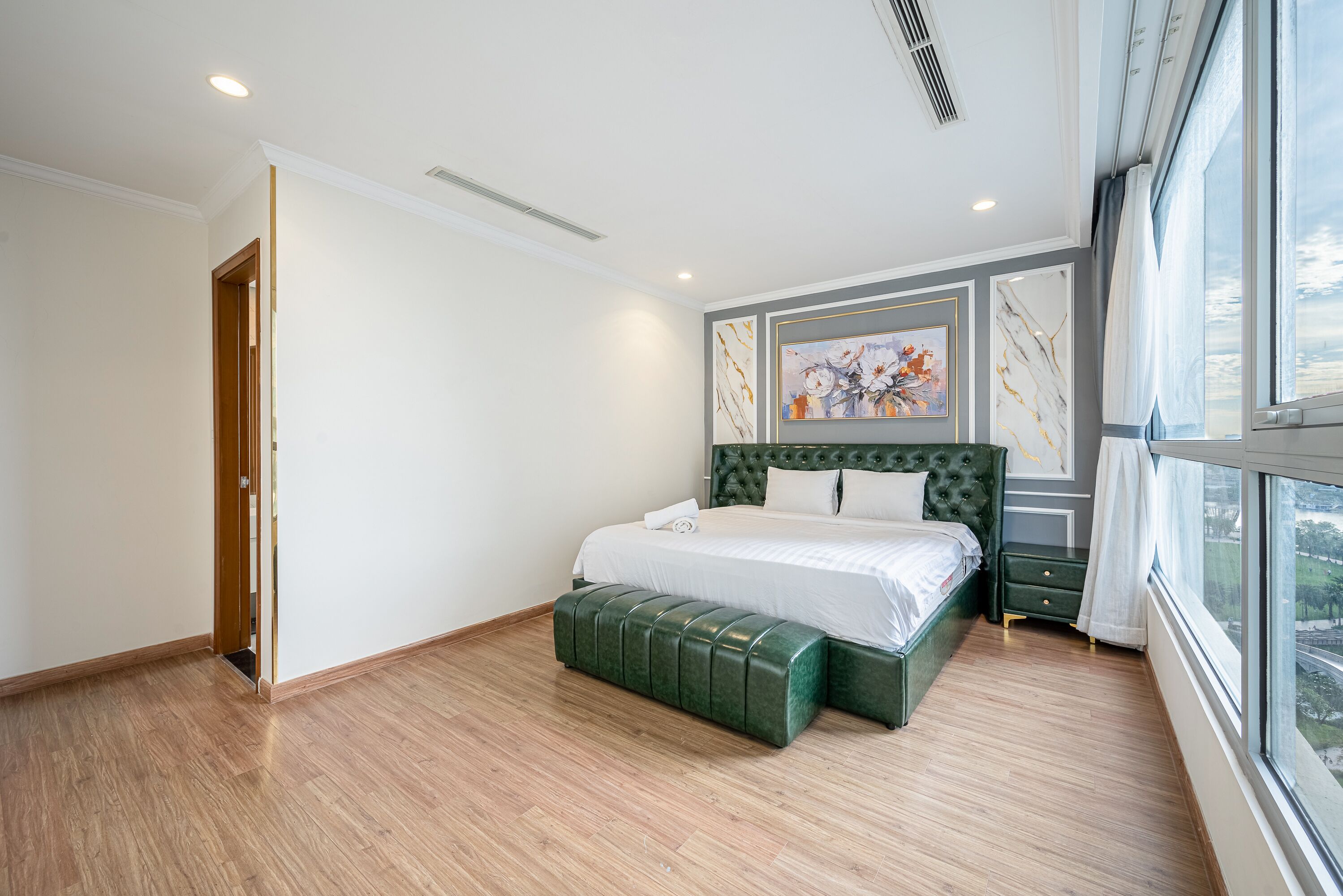 Un dormitorio con una cama grande, un banco verde y una ventana con vista a la ciudad.