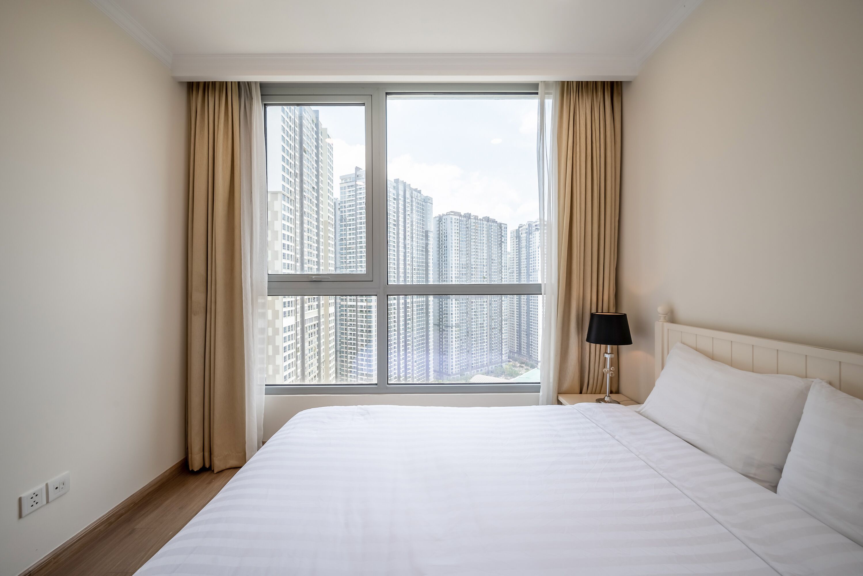 Habitación de hotel con una cama grande, una mesita de noche con lámpara y ventana con vista a la ciudad.