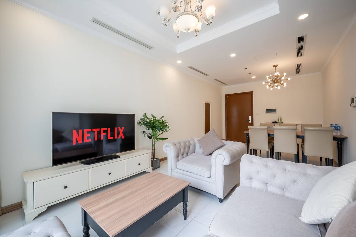 A modern living room with a flat-screen TV, a sofa, a coffee table, and a dining area in the background.