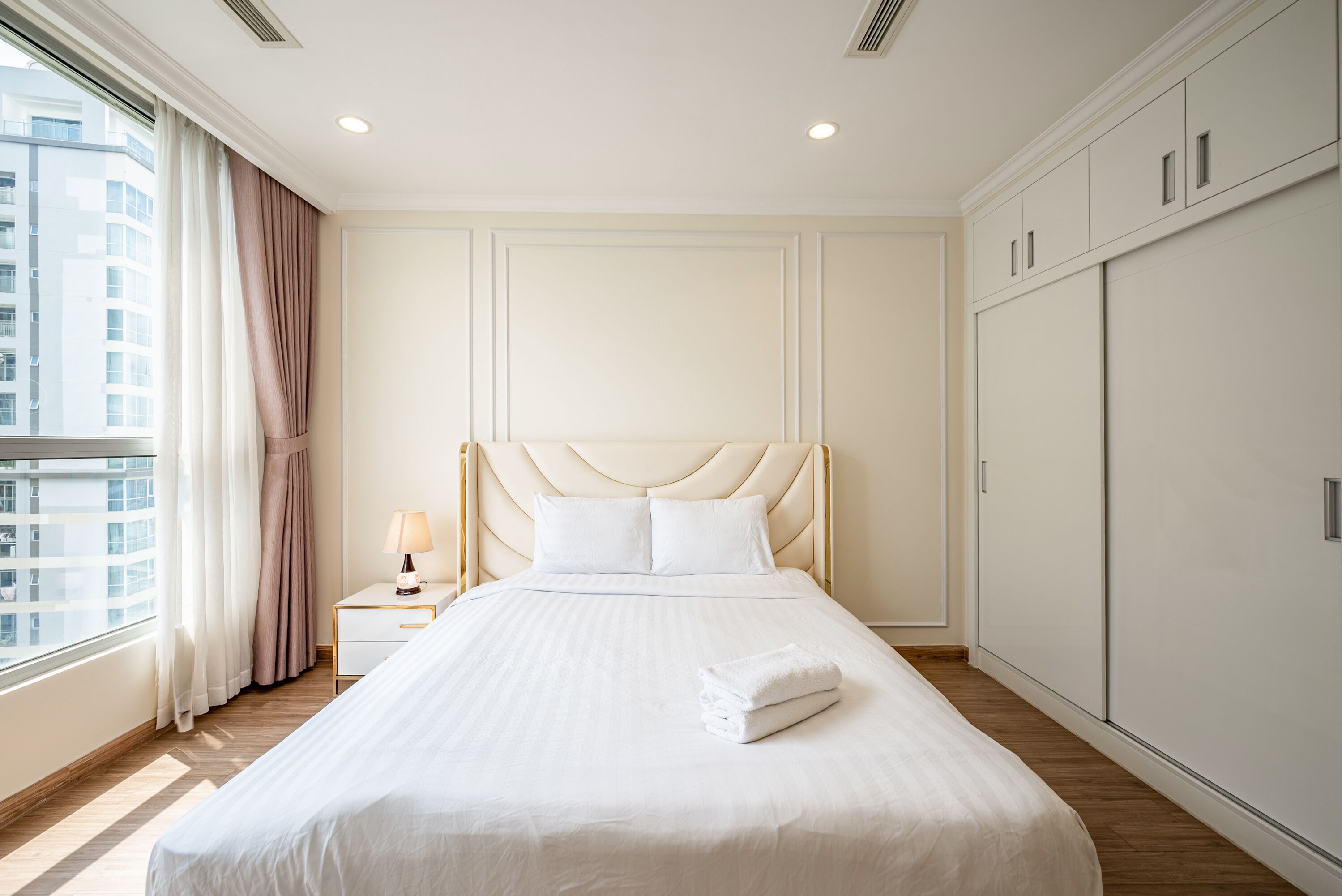 A hotel room with a large bed, a nightstand with a lamp, a wardrobe, and a window with curtains.