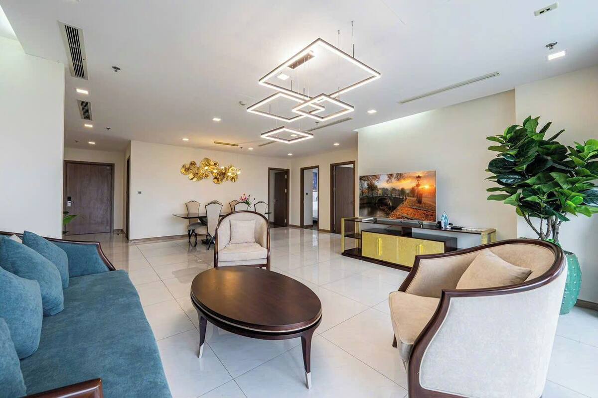A modern living room with a sofa, armchairs, a coffee table, and a TV. There is a dining area with chairs and a table, and a large plant.