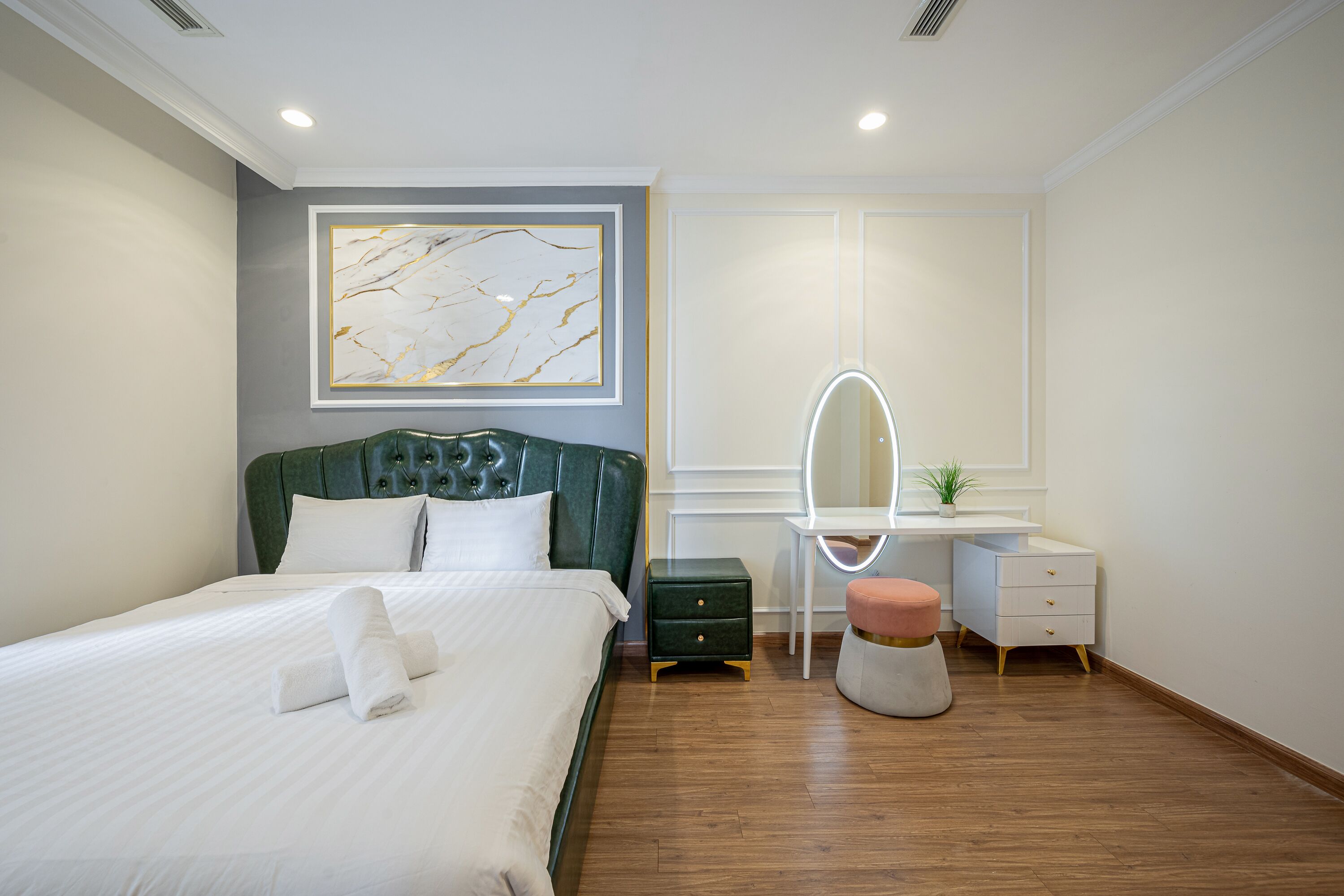 A neatly made bed with white linens, a green upholstered headboard, a small bedside table, a white dresser with drawers, a round mirror, and a potted plant.