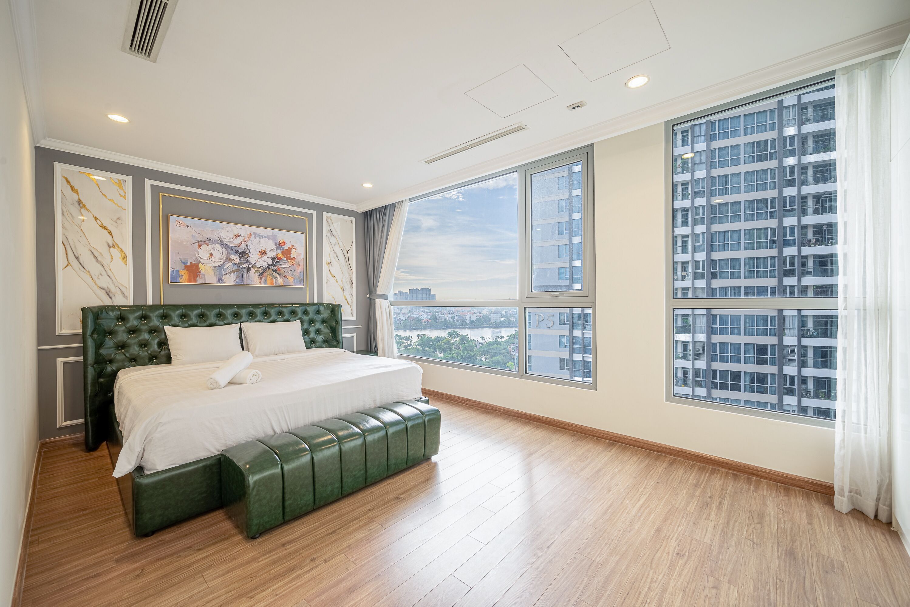 Un dormitorio amplio con una cama grande, un banco tapizado de color verde y vistas a la ciudad a través de amplias ventanas.