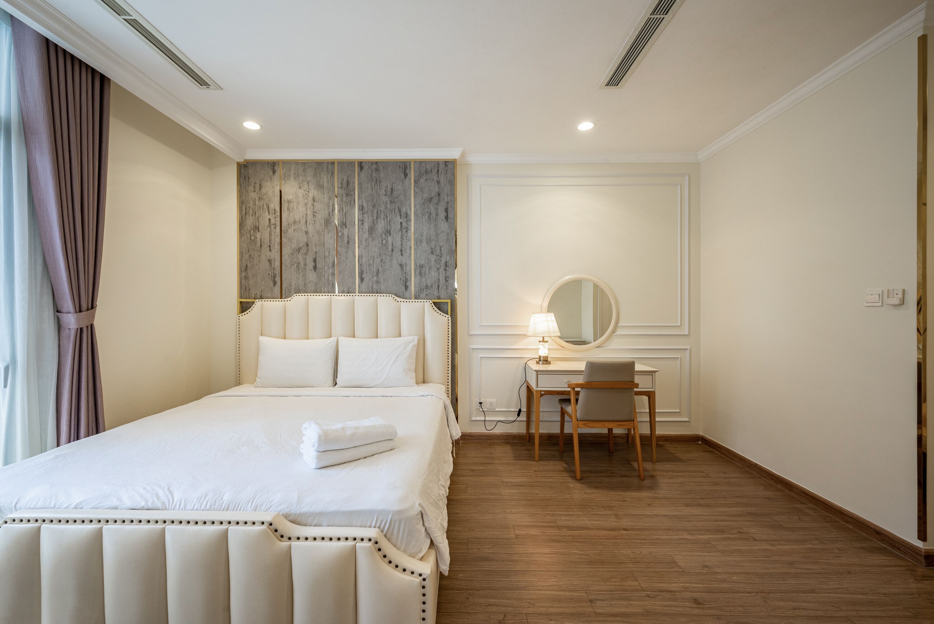 A hotel room with a large bed, a desk with a chair, a round mirror, and a wooden headboard.