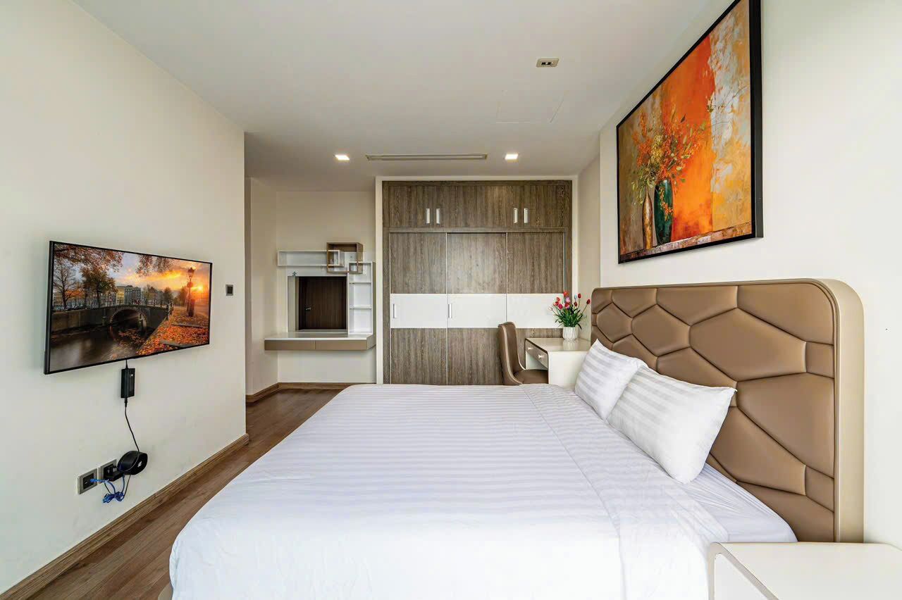 A hotel room with a large bed, a TV, a painting, and a kitchenette in the background.