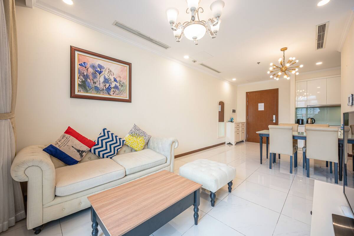 A modern living room with a sofa, ottoman, and a wooden coffee table. There is a dining area with a table and chairs, and a kitchen with cabinets and appliances.