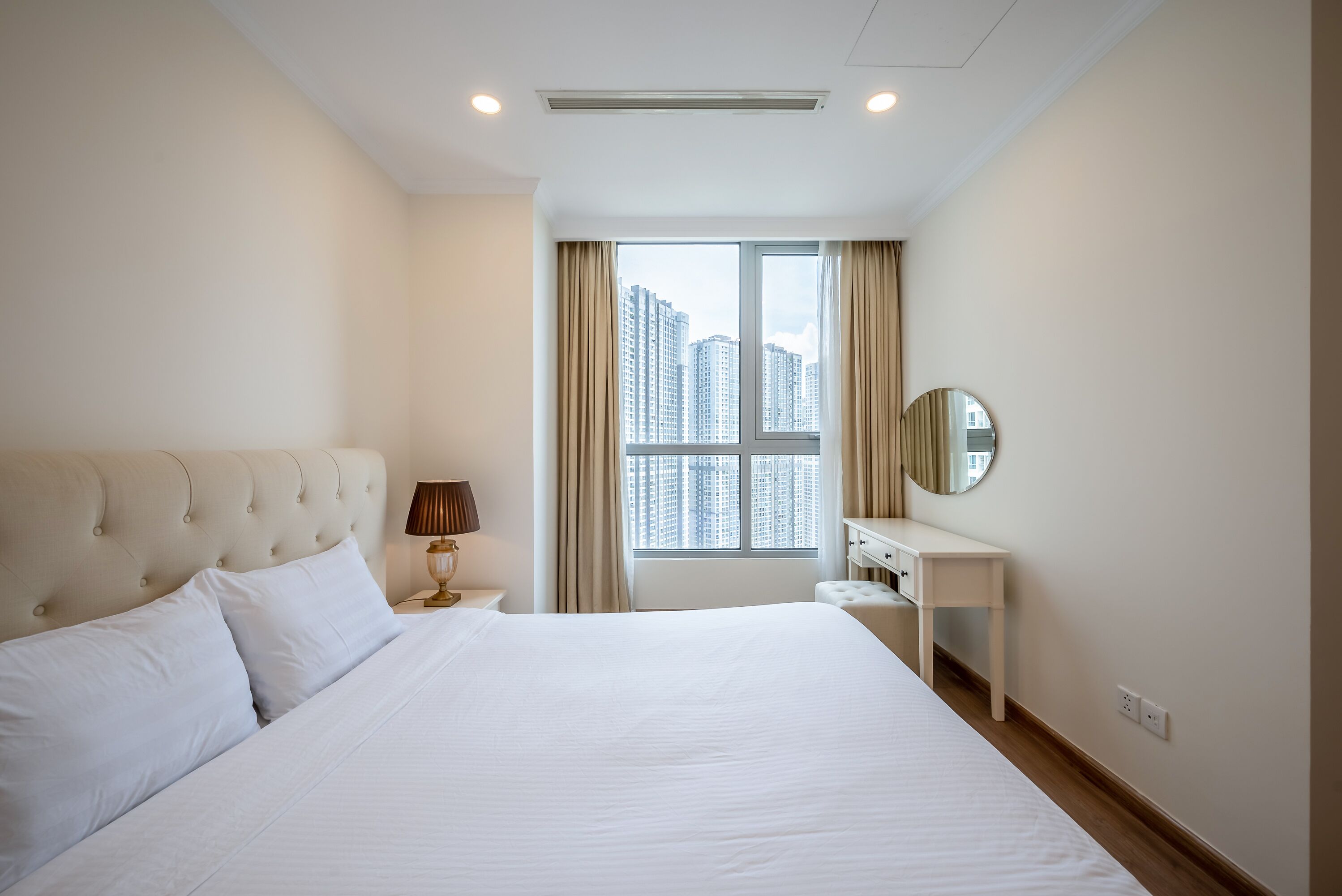 Habitación de hotel con una cama grande, una mesita de noche con lámpara, un escritorio pequeño, un espejo y una ventana con vista a la ciudad.