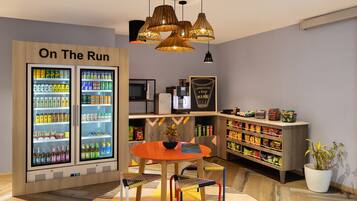 A modern room with a "On The Run" refrigerator stocked with beverages, a coffee machine, a dining table with chairs, and a bookshelf with snacks.