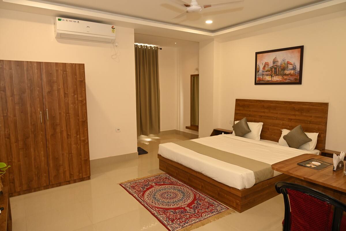 A hotel room with a wooden bed, a desk with a chair, a red patterned rug, a painting on the wall, and an air conditioning unit.