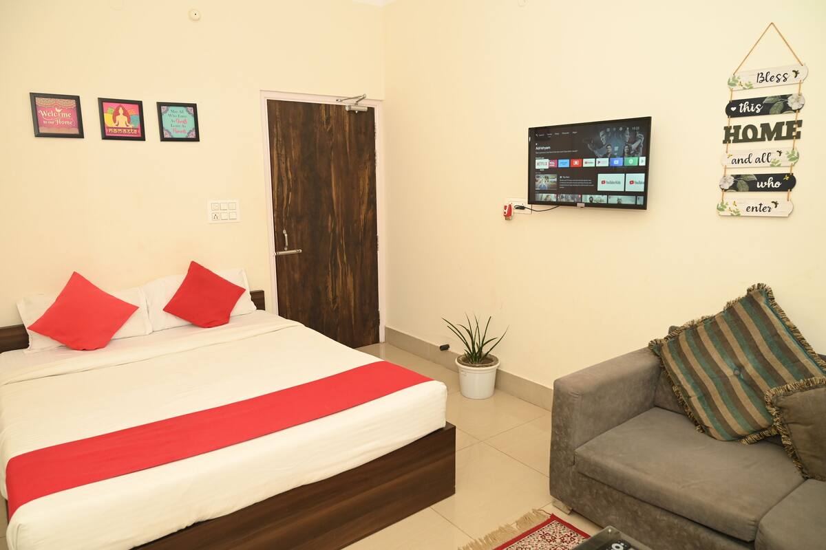 A bedroom with a bed, a grey sofa, a TV, and a door.