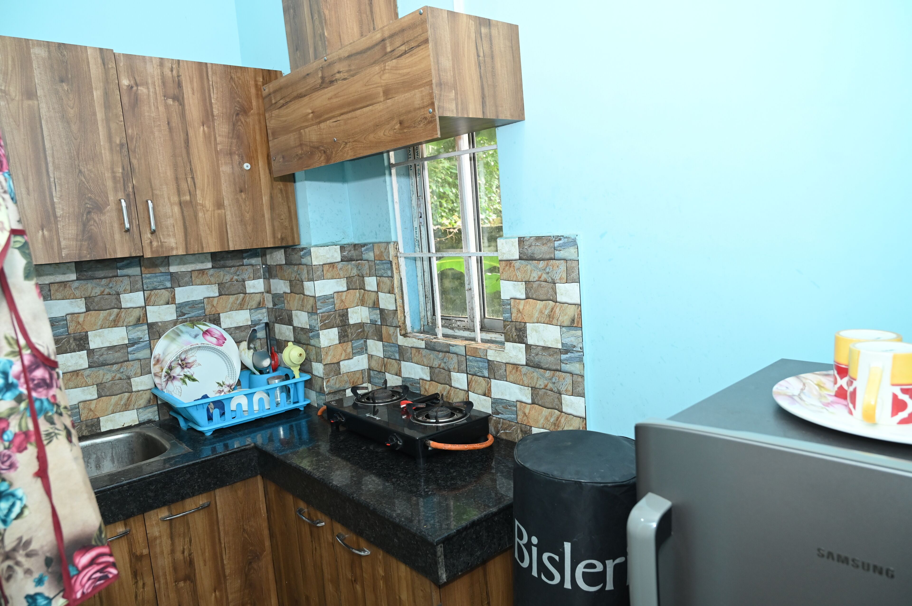 Uma cozinha com armários de madeira, painel de azulejos, janela, geladeira Samsung e escorredor de louça.