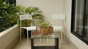 Un balcón con dos sillas blancas, una cesta de mimbre para bicicleta y plantas en macetas.