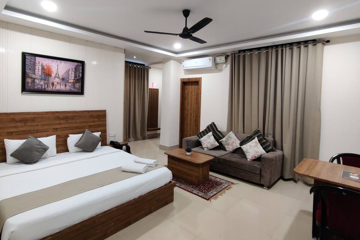 A modern hotel room with a large bed, a sofa, a dining table, and a ceiling fan.