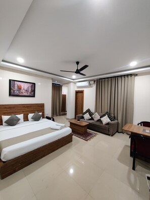 A modern hotel room with a large bed, a sofa, a dining table, and a ceiling fan.