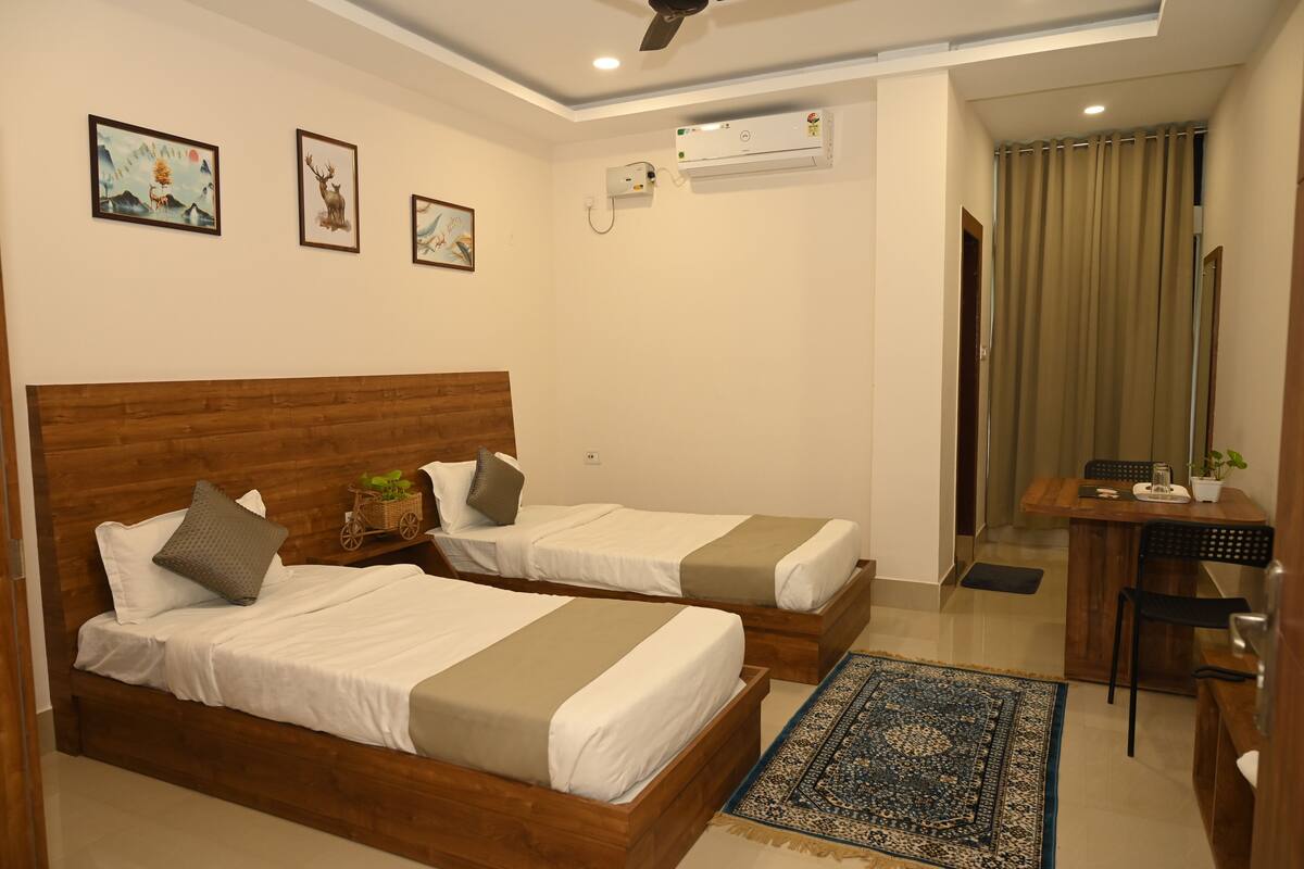 A hotel room with two beds, a desk, and a chair. There are wall-mounted air conditioning units and framed pictures on the wall.