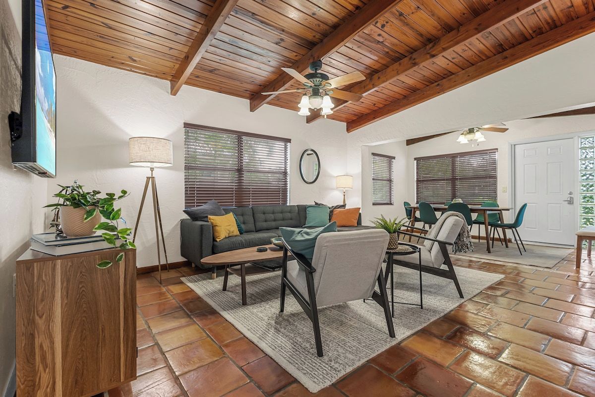 A spacious living area with a wooden ceiling, a sofa set, a dining table with chairs, and a television mounted on the wall.