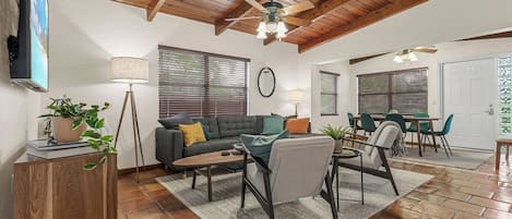 A spacious living area with a wooden ceiling, a sofa set, a dining table with chairs, and a television mounted on the wall.