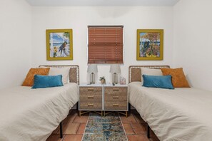 A bedroom with two beds, a nightstand with drawers, a window with blinds, and two framed pictures on the wall.