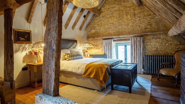 A rustic bedroom with exposed wooden beams, a stone wall, a bed with a yellow throw, a wooden nightstand, and a chair.
