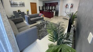 Een moderne woonkamer met een grijze bank, een glazen salontafel en een eethoek met een tafel en stoelen.