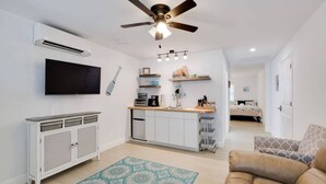 A modern living room with a ceiling fan, a flat-screen TV, a kitchen area with a countertop, and a patterned rug.