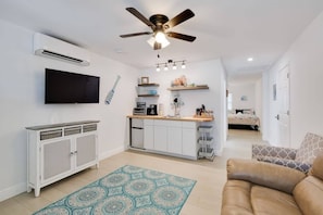 A modern living room with a ceiling fan, a flat-screen TV, a kitchen area with a countertop, and a patterned rug.