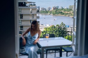 Un balcon avec une table et un verre de vin, donnant sur une rivière où naviguent des bateaux, avec une vue sur la ville en arrière-plan.