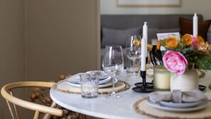 Una mesa redonda para comer con un tablero de mármol blanco, rodeada de sillas de mimbre. Hay platos, vasos y un florero con flores sobre la mesa.