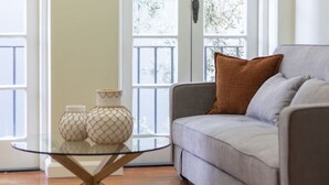 Una sala de estar con un sofá gris, una mesa de centro de vidrio con dos jarrones estampados y una puerta blanca que da a vistas de árboles.