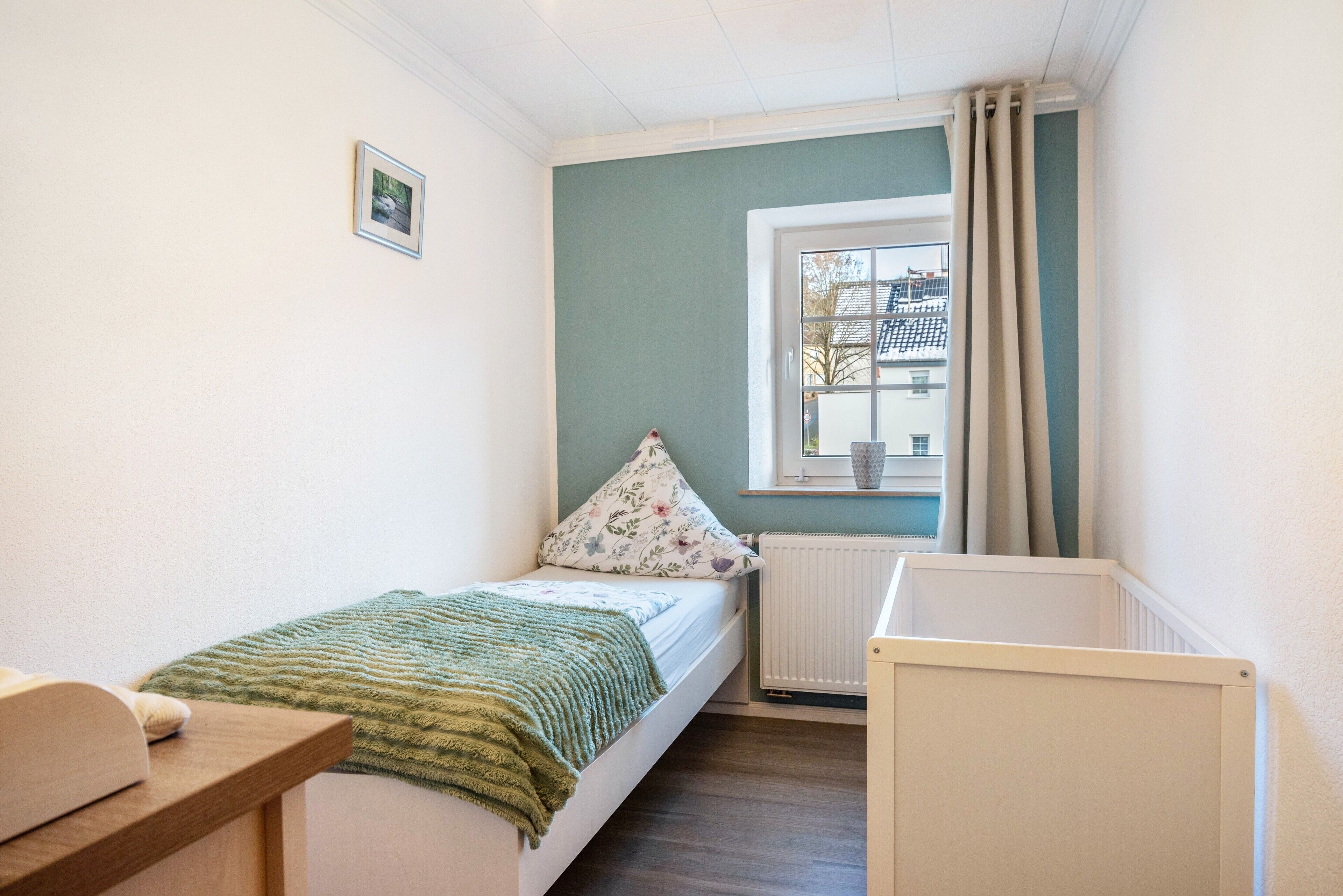 A small, tidy room with a single bed, a window with curtains, a radiator, and a small table.