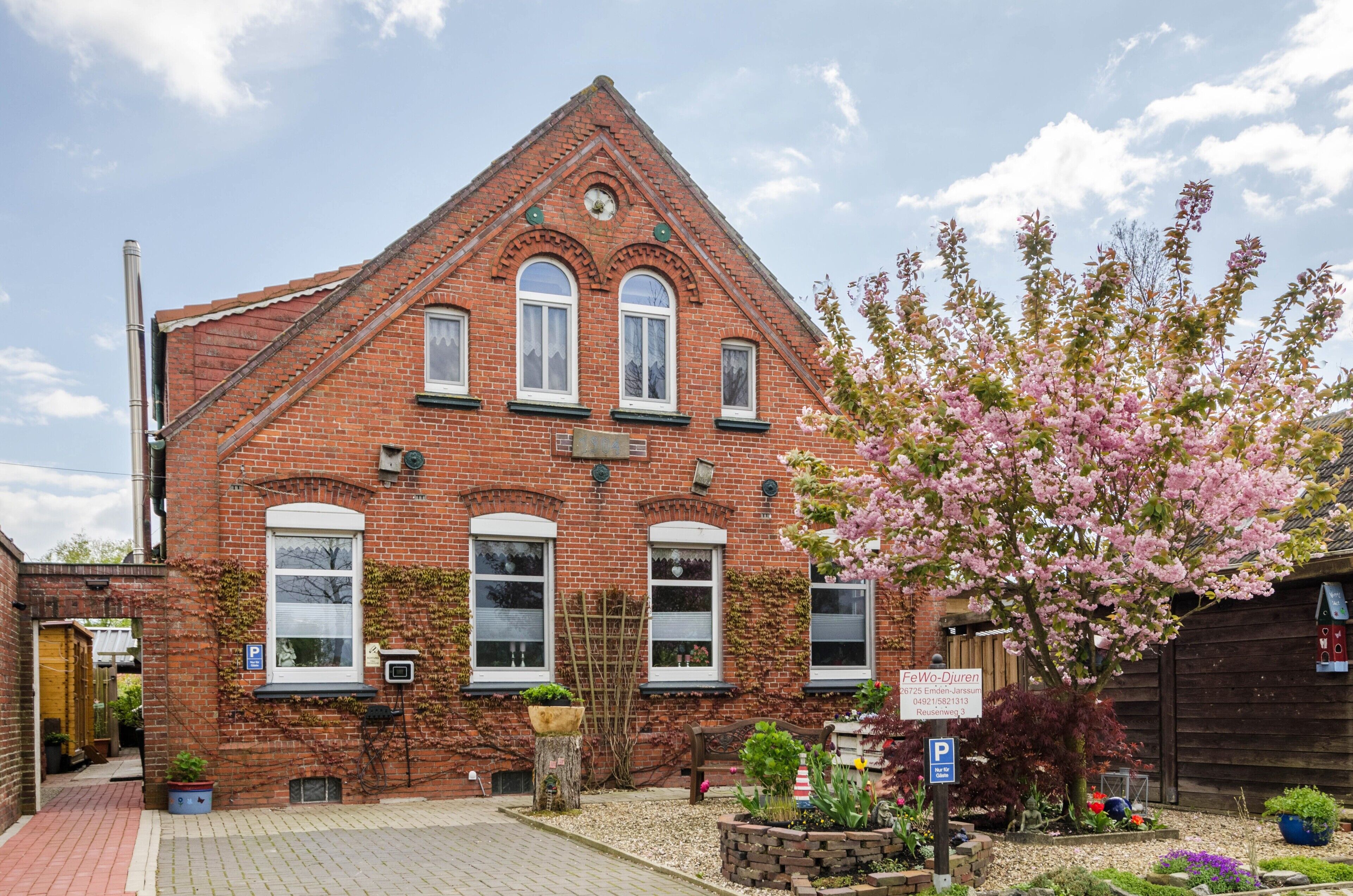 A red brick building with a triangular roof, a clock, and a sign with a name and address.