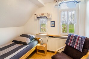 A small, well-lit room with a single bed, a chair, a desk with a lamp, and a window with a view of greenery. - Apartment 'Djuren' with Private Terrace, Shared Garden, and Wi-Fi (Niedersachsen - Emden)
