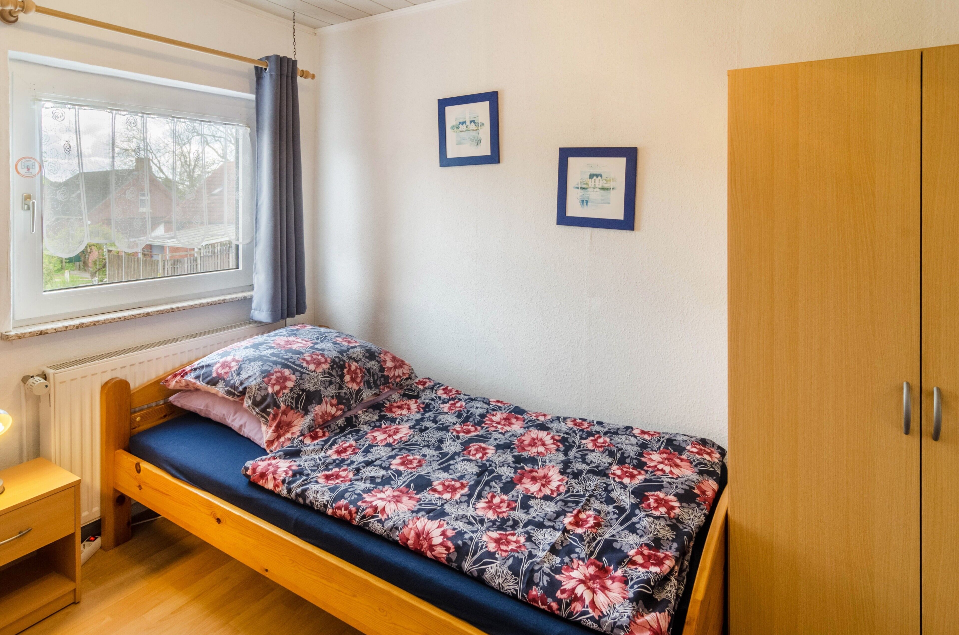 Yhden hengen sänky, jossa on kukallinen päiväpeite, puinen vaatekaappi, yöpöytä ja ikkuna, jossa on verho.