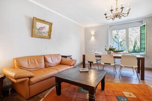 A living room with a brown leather sofa, a wooden coffee table, a dining area with a table and chairs, a chandelier, and a window with a view of greenery.