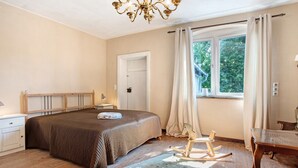 A bedroom with a large bed, a wooden rocking chair, a small table, and a window with curtains.