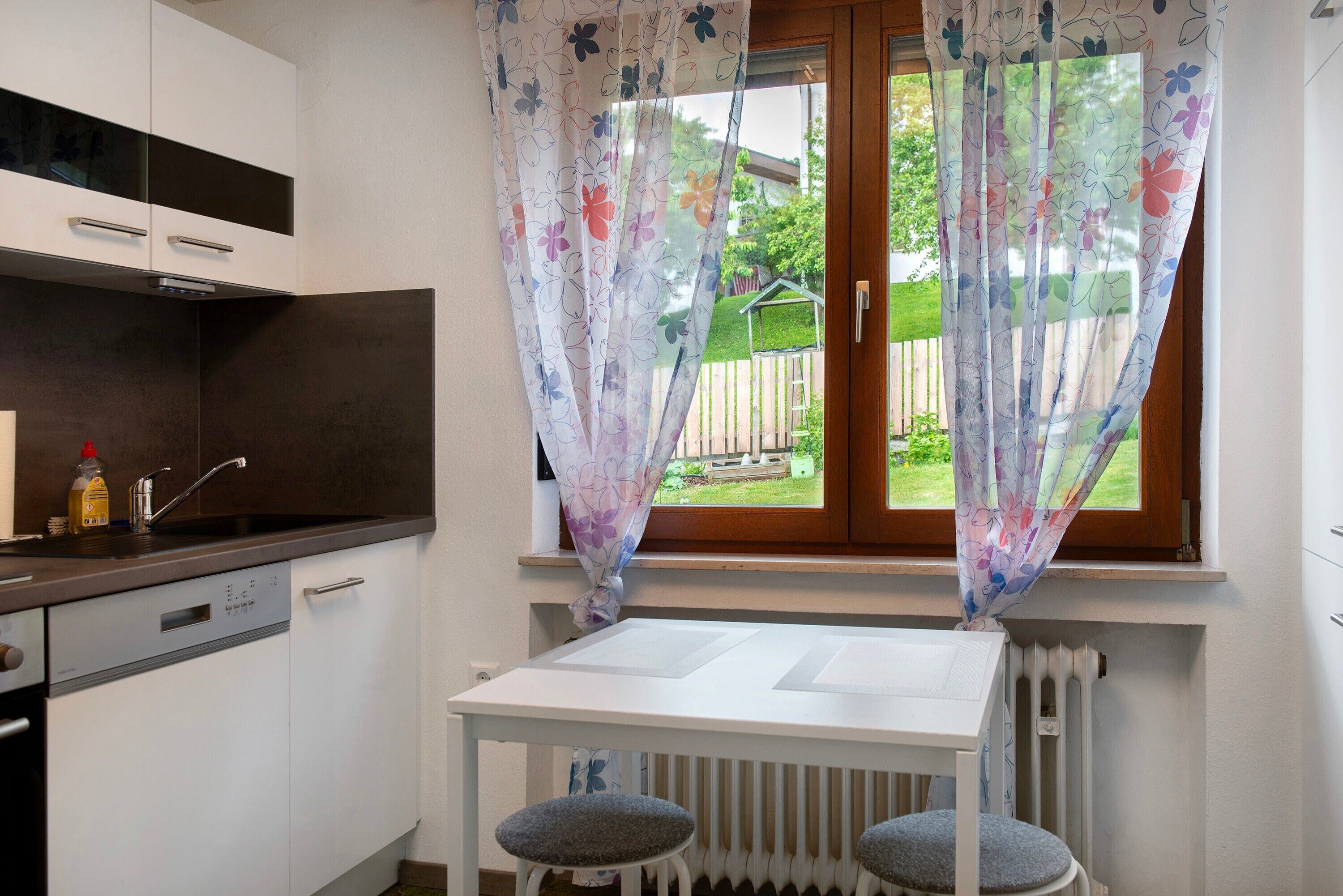 Una cocina con encimera blanca, lavavajillas y una ventana con cortinas florales.