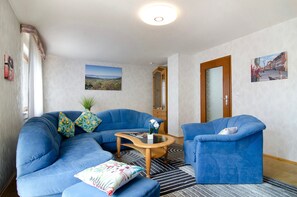 A living room with a blue sofa, a matching armchair, a coffee table, and a rug. There is a door with a mirror, a painting on the wall, and a window with curtains.