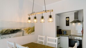 A dining area with a wooden table and white chairs, a kitchen with a stove and cabinets, and a beach-themed wall art. - Holiday Apartment 'Ferienhaus Deichblick Nessmersiel' with Private Terrace, Garden & Wi-Fi (Dornum)