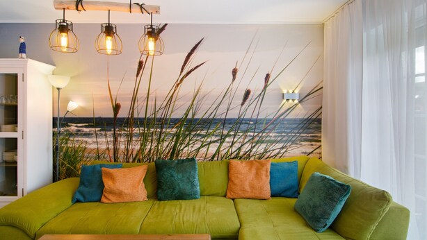 A green sofa with colorful cushions, a wooden table, and a view of the beach through a window. - Holiday Apartment 'Ferienhaus Deichblick Nessmersiel' with Private Terrace, Garden & Wi-Fi (Dornum)