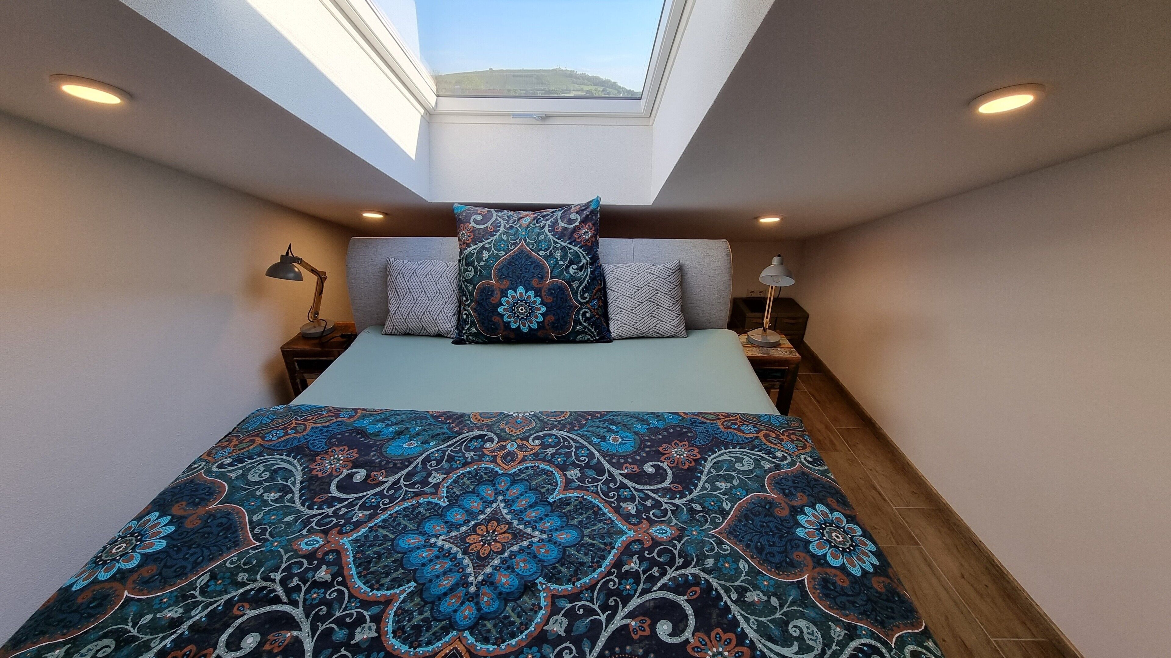 A bedroom with a skylight, a bed with patterned bedding, two bedside lamps, and a view of a landscape through the skylight.