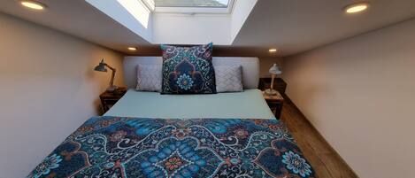 A bedroom with a skylight, a bed with patterned bedding, two bedside lamps, and a view of a landscape through the skylight.