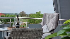 A balcony with a wicker chair, a table with a bottle of wine and glasses, and a view of greenery and buildings.