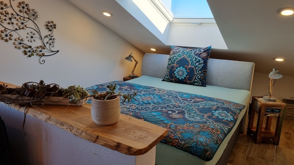 A bedroom with a skylight, a bed with patterned bedding, a bedside table with a lamp, and a decorative wall piece.