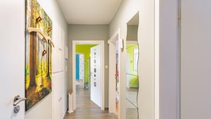 A hallway with a door on the left, a painting on the wall, and a door with a green door frame in the background.