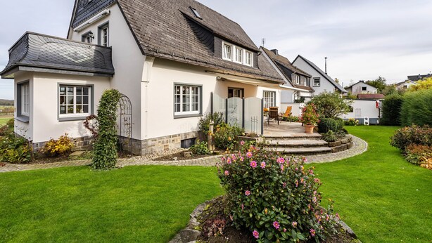 A two-story house with a sloped roof, a front porch, and a well-maintained garden with flowers and shrubs.